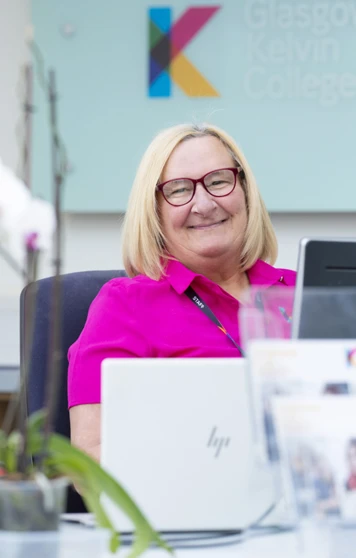 Receptionist smiling at her desk in the Glasgow Kelvin College West Campus, with a Glasgow Kelvin College sign in the background and orchids in the foreground. Receptionist smiling at her desk in the Glasgow Kelvin College West Campus, with a Glasgow Kelvin College sign in the background and orchids in the foreground.